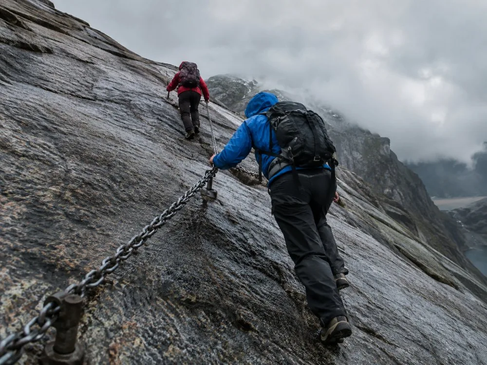 Hikers holding the fixed chains bolted along a steep bare slab on the Kjerag trail